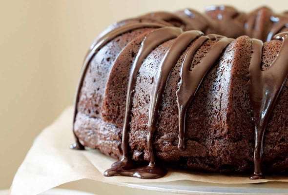 A chocolate glazed chocolate bundt cake sitting atop a white cake stand with a dish of glaze on the side