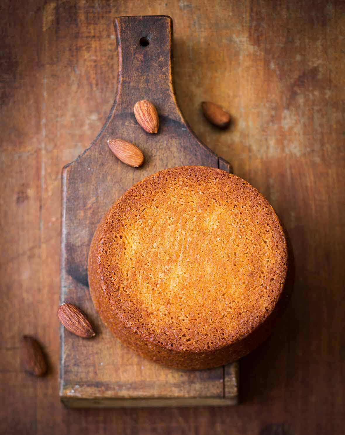 A round flourless almond cake on a wooden baking paddle with almonds scattered around it