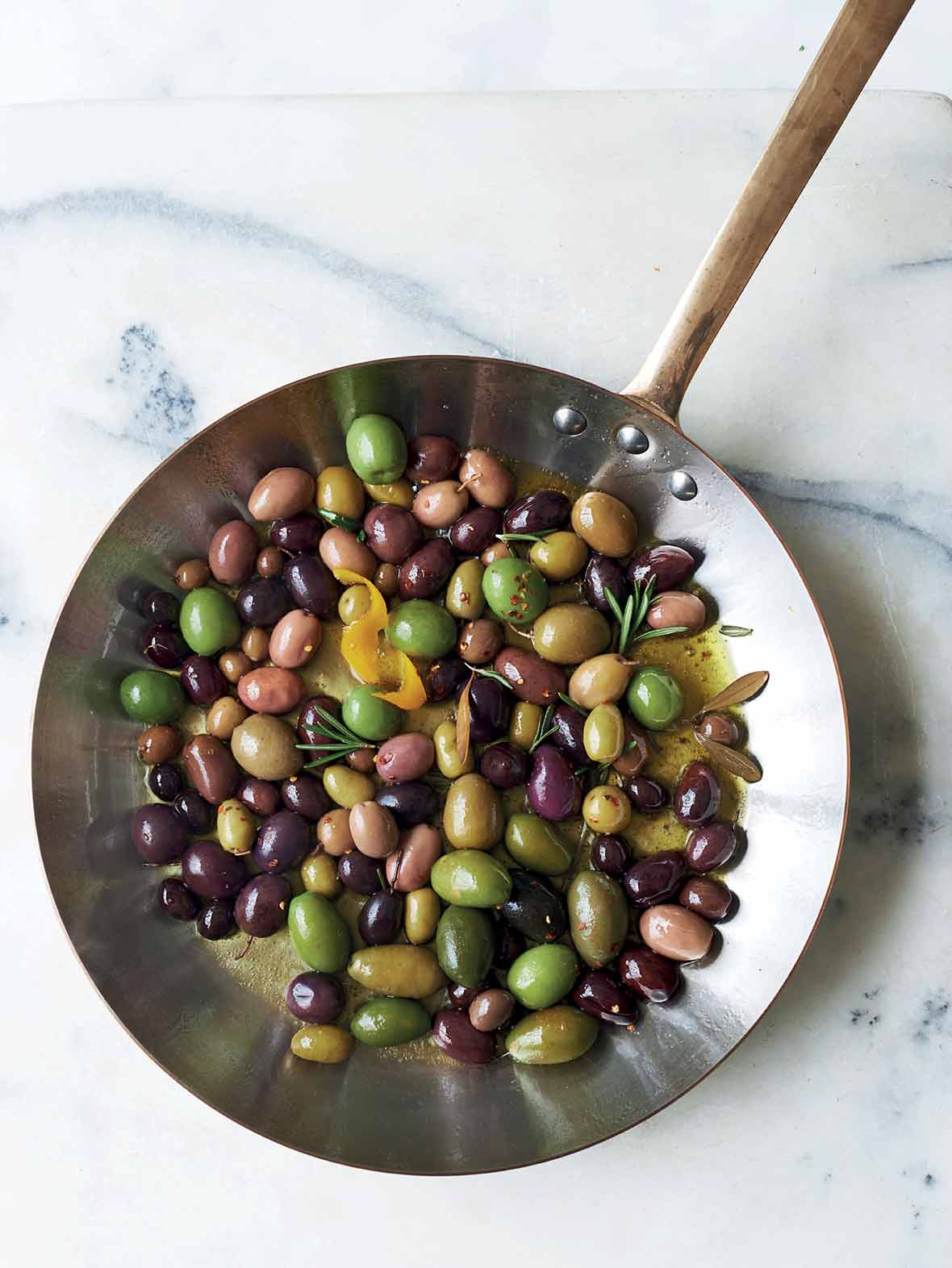 A metal skillet filled with warm olives, rosemary, and lemon zest.
