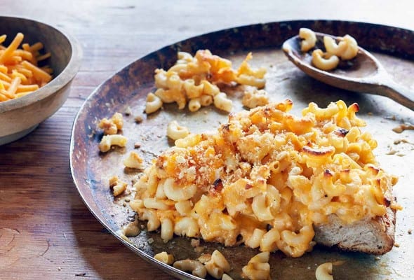 One slice of mac and cheese pizza on a rimmed pizza pan with a wooden spoon resting on the sheet.
