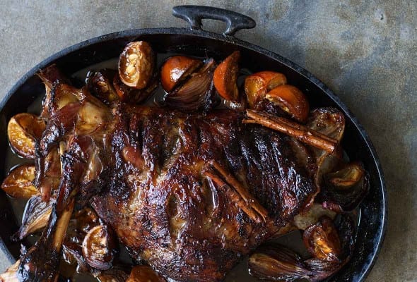 A cast-iron cooking dish filled with roasted lamb shoulder and orange halves, topped with cinnamon sticks