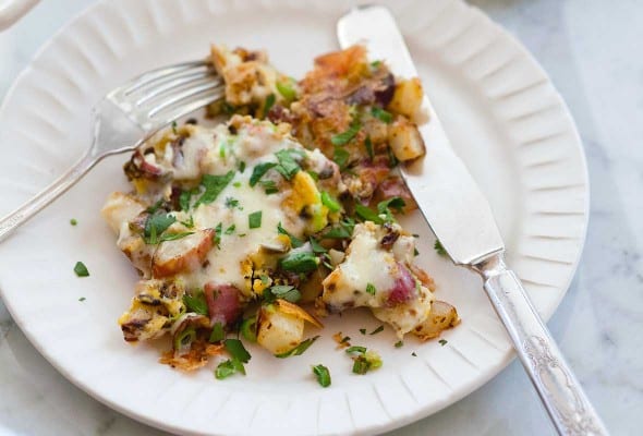 A white plate of fried potatoes with eggs, covered in cheese with a skillet in the background and a fork and knife on the plate.