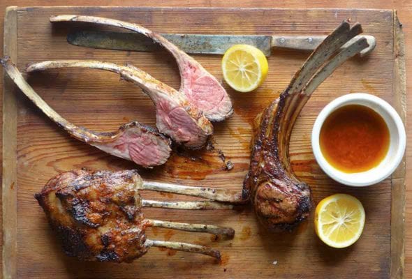 A cutting board with lamb chops, some in racks, some sliced, along jus and lemon halves