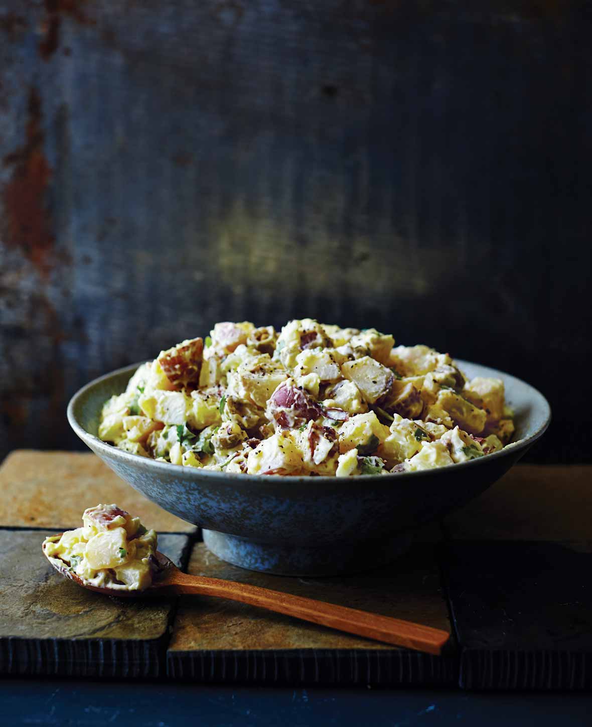 A blue bowl filled with Southern potato salad and a wooden spoonful of salad lying beside it.