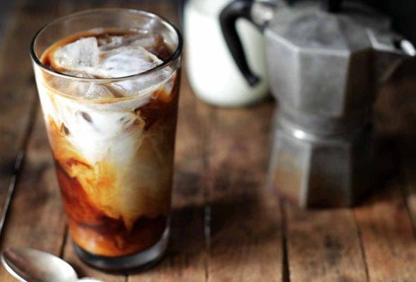 A glass of cold brew coffee with a French press and jar of cream in the background.