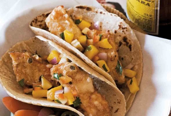 A basket containing two Baja-style tempura fish tacos and some pickled vegetables, with a bottle of Pacifico on the side.