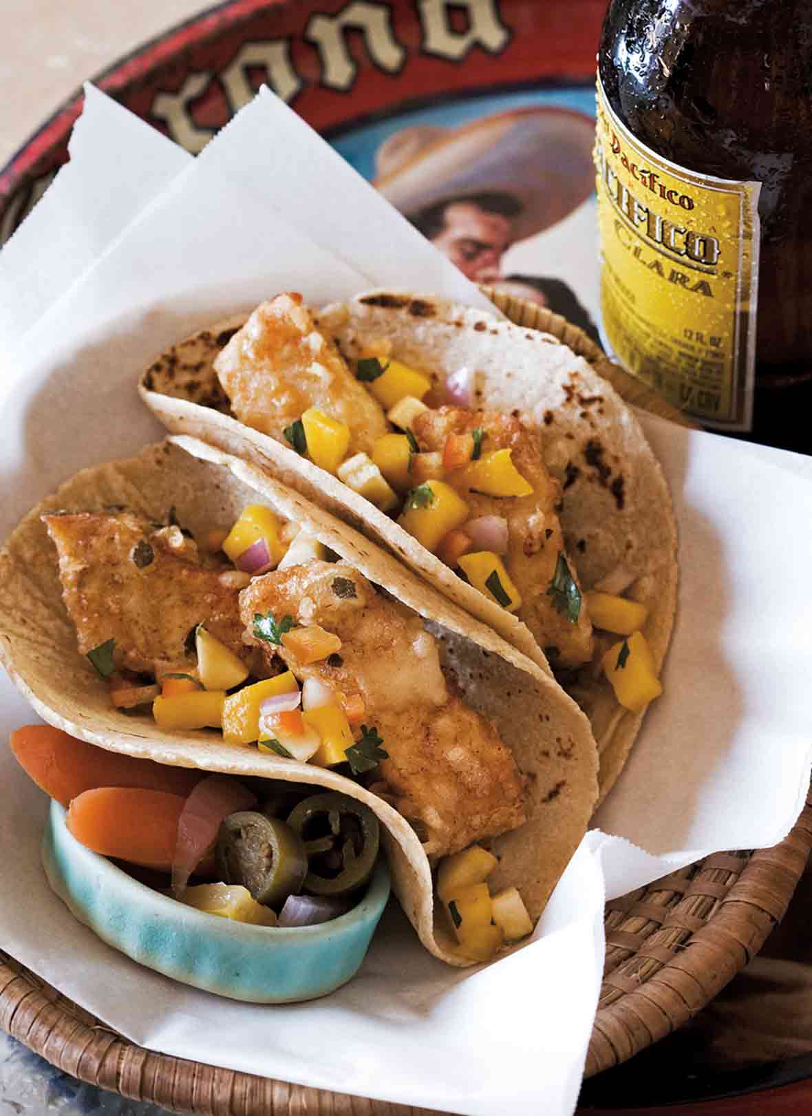 A basket containing two Baja-style tempura fish tacos and some pickled vegetables, with a bottle of Pacifico on the side.