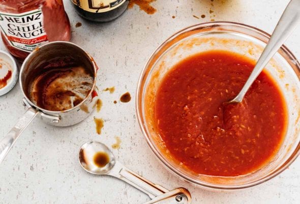 A glass bowl of homemade cocktail sauce with a spoon resting inside and bottles of chili sauce, hot sauce, and Worcestershire sauce in the background.