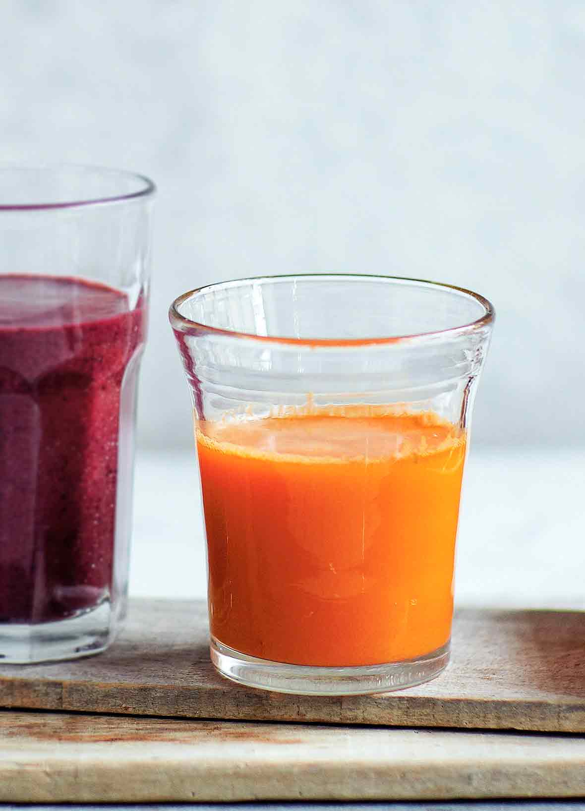 A glass of carrot ginger juice, and another glass of blueberry spinach smoothie on a wooden plank.