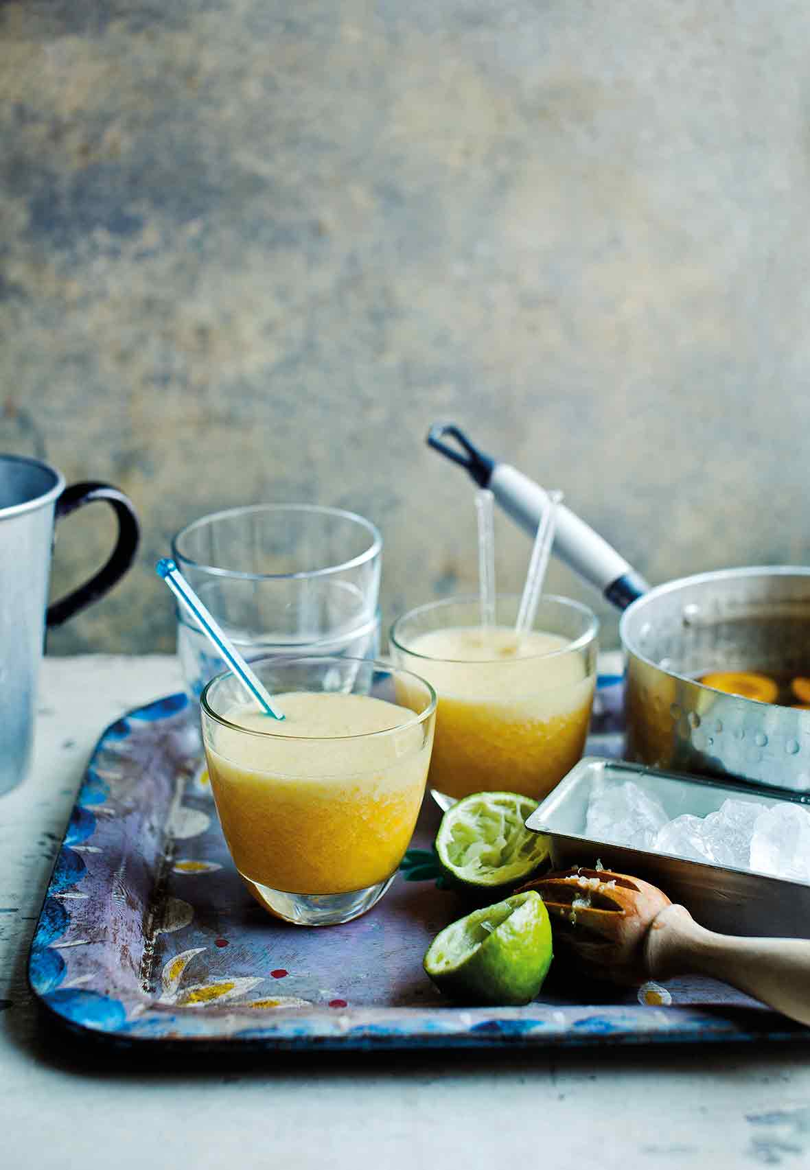 A metal serving tray with two apricot daiquiris with stir sticks, a juiced lemon, and a pot with poached apricots.