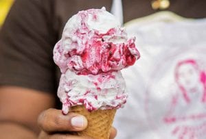 A person holding a sugar cone topped with two scoops of blackberry ice cream.
