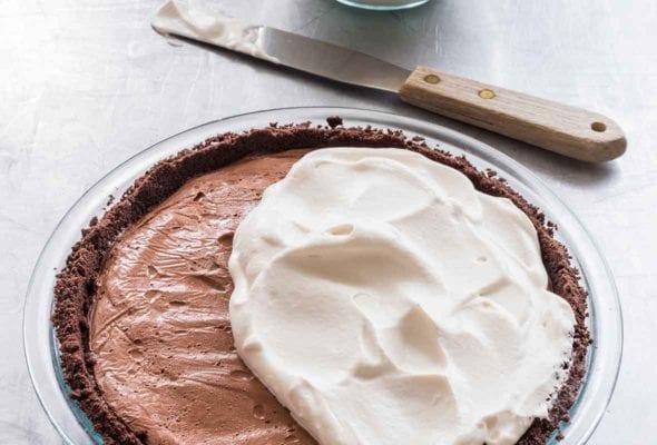 A dark chocolate cream pie half covered with whipping cream with a bowl of whipping cream and a spatula resting beside the pie.