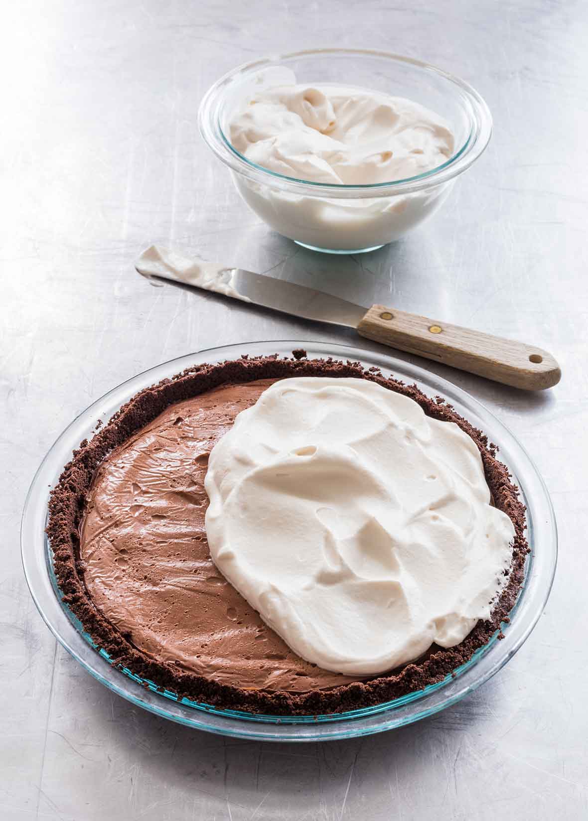 A dark chocolate cream pie half covered with whipping cream with a bowl of whipping cream and a spatula resting beside the pie.