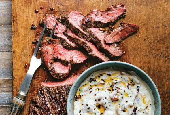 Sliced grilled hanger steak with raita in a bowl beside the steak on a wooden cutting board.