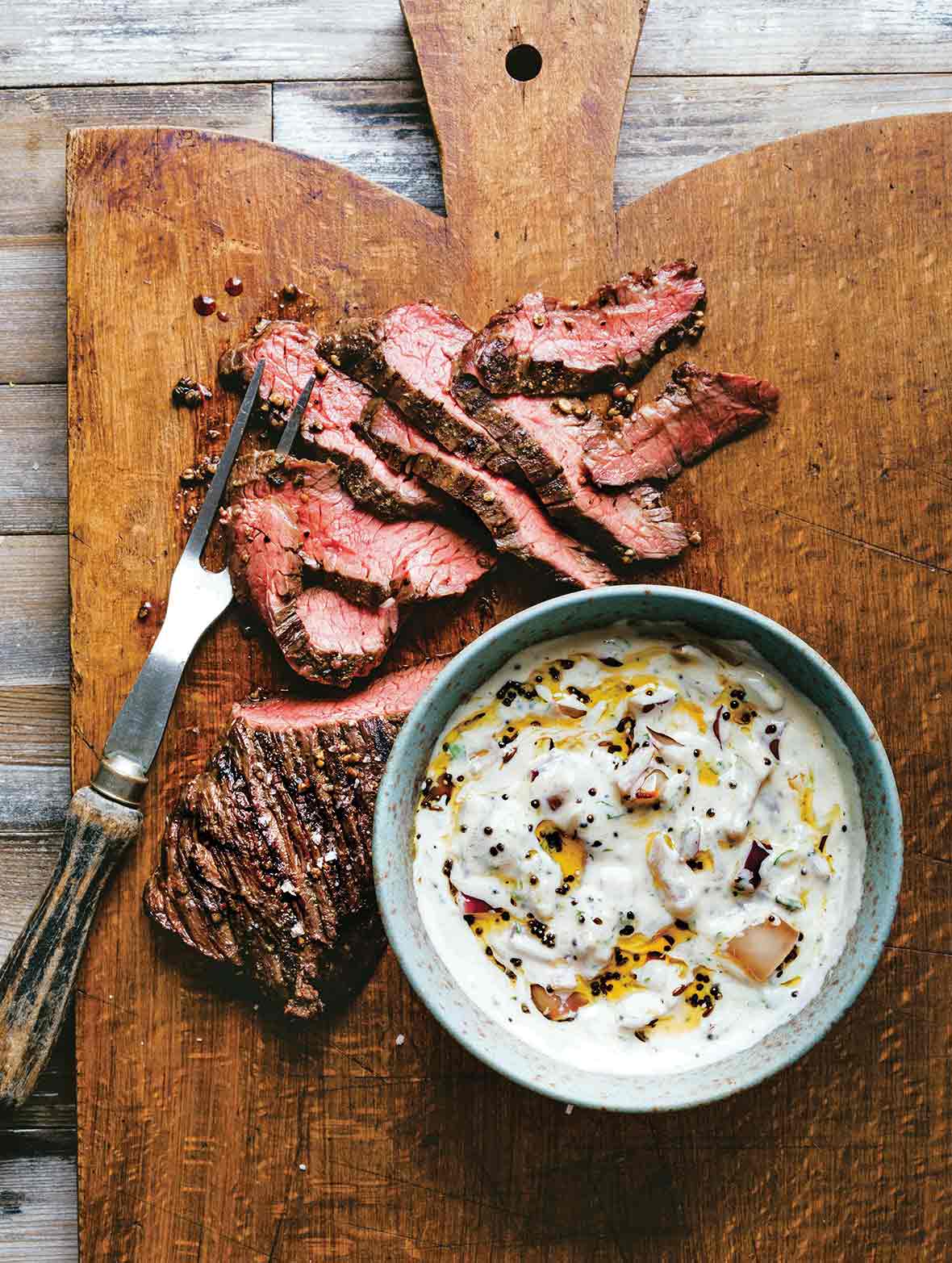 Sliced grilled hanger steak with raita in a bowl beside the steak on a wooden cutting board.