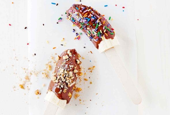 Two chocolate banana pops, one covered in sprinkles and the other in chopped nuts on a piece of parchment.