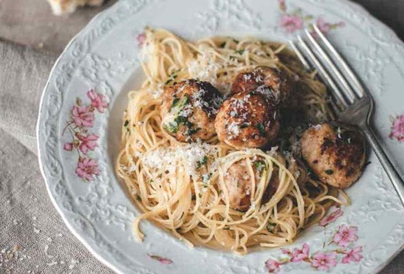 A patterned bowl filled with turkey meatballs with angel hair pasta, and a piece of bread and glass of white wine next to the bowl.