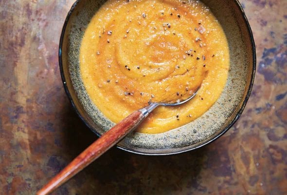 A bowl of curried sweet potato and lentil soup, sprinkled with black pepper, with a spoon resting inside the bowl.