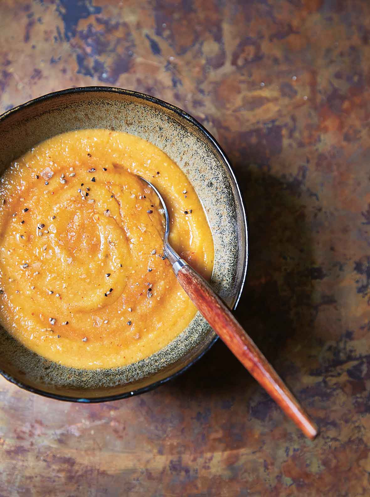 A bowl of curried sweet potato and lentil soup, sprinkled with black pepper, with a spoon resting inside the bowl.