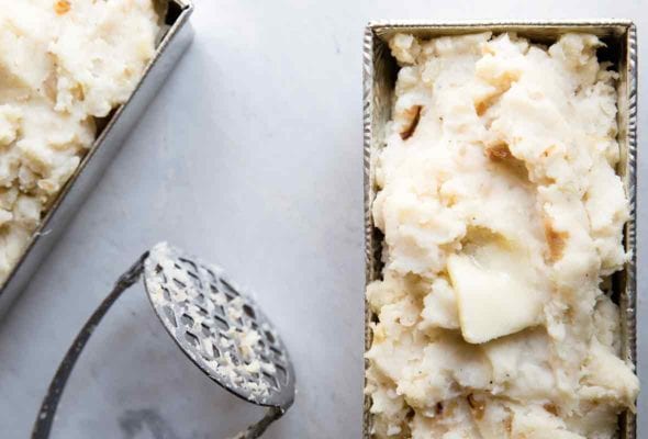 Two metal loaf pans filled with roasted garlic mashed potatoes with a masher resting in between them.