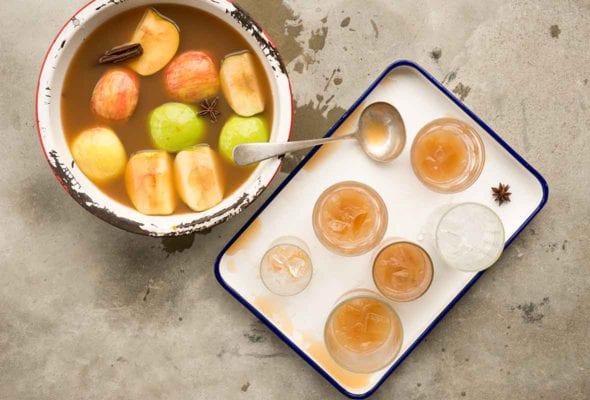 A chipped pot filled with apple pie moonshine, apple wedges and whole spices with a tray of glasses filled with the moonshine beside it.