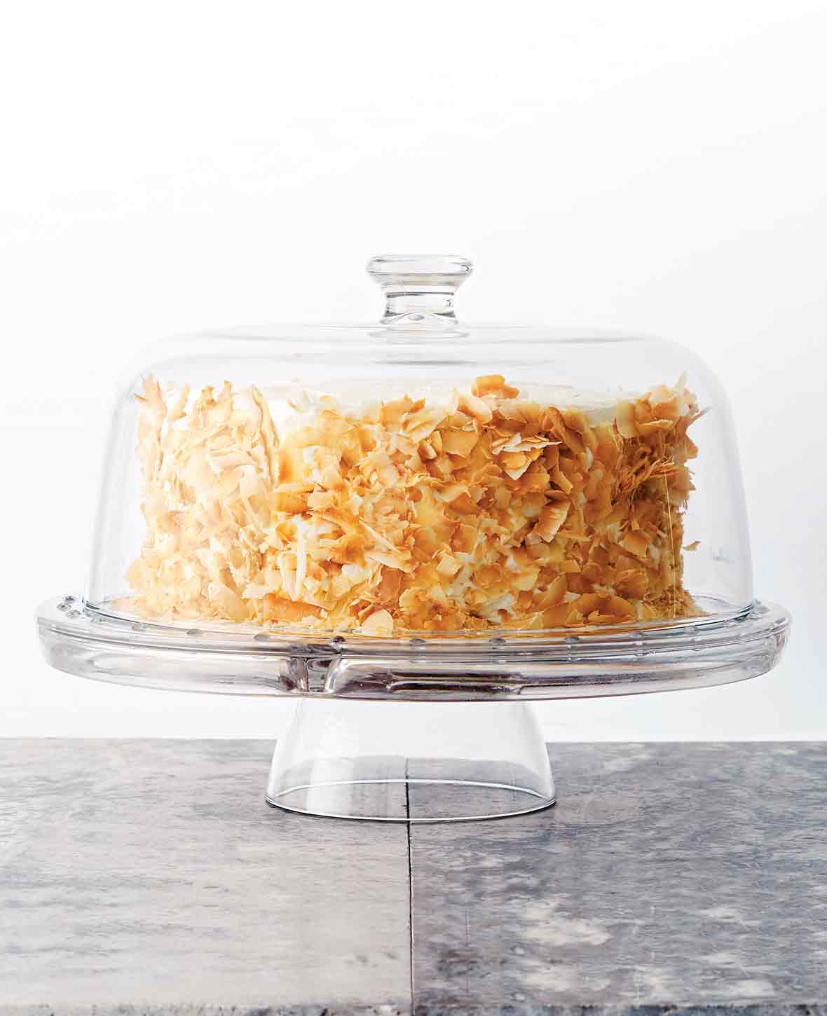 A coconut carrot cake with cream cheese frosting and toasted coconut on the sides on a glass cake platter with a glass domed lid.