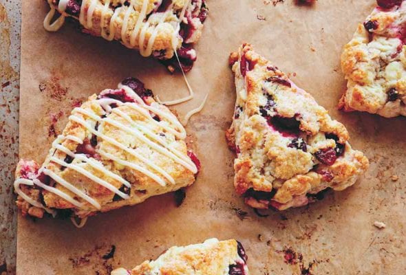 Five cranberry scones with two drizzled with white chocolate glaze on a parchment-lined rimmed baking sheet.