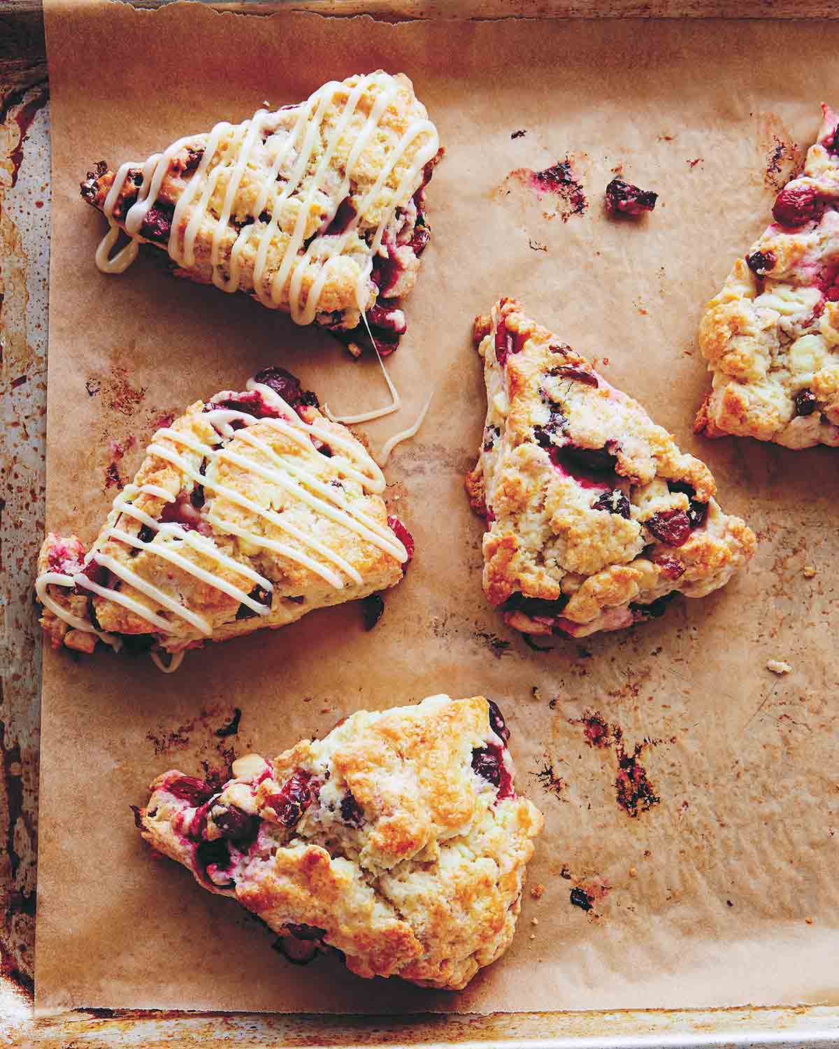 Five cranberry scones with two drizzled with white chocolate glaze on a parchment-lined rimmed baking sheet.