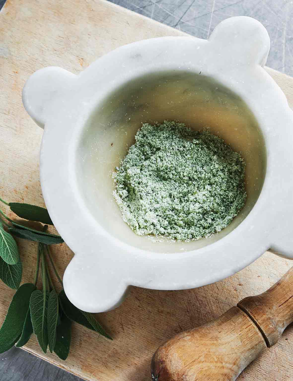 A mortar partially filled with herb salt and a pestle and two sprigs of fresh sage leaves lying beside it.