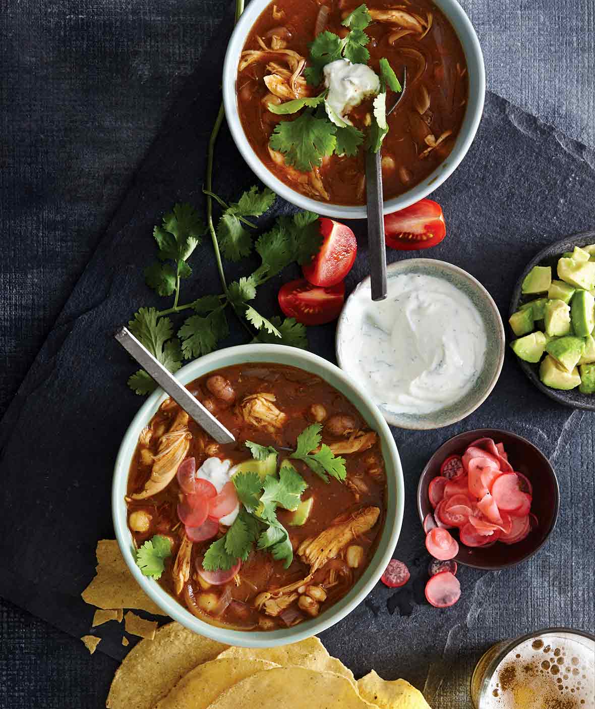 Two bowls of chicken posole, with a bowl of sour cream, some tomato wedges, cilantro, sliced radish, and tortilla shells scattered around them.