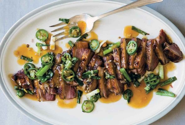 A white oval platter filled with sliced duck à l’orange, topped with sliced scallions and jalapenos, and drizzled with sauce.