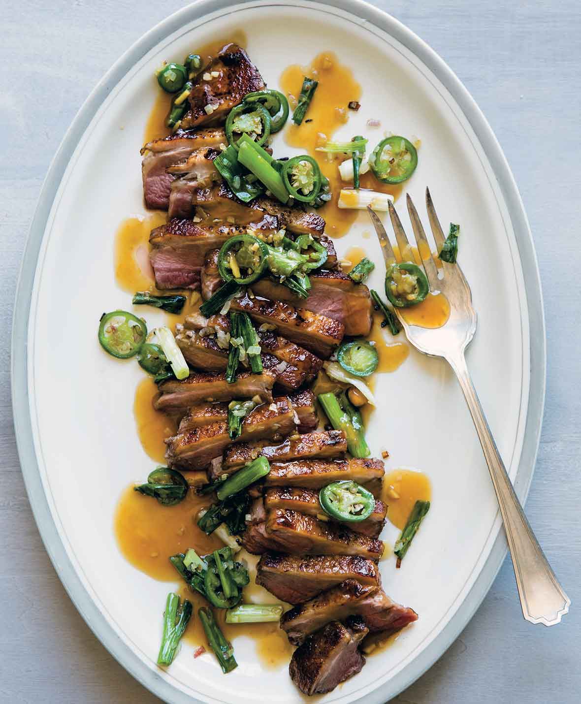 A white oval platter filled with sliced duck à l’orange, topped with sliced scallions and jalapenos, and drizzled with sauce.