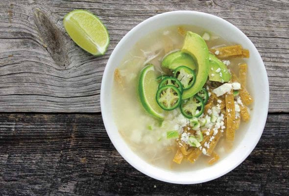 A white bowl filled with broth, avocado slices, lime wedges, cheese, and tortillas strips