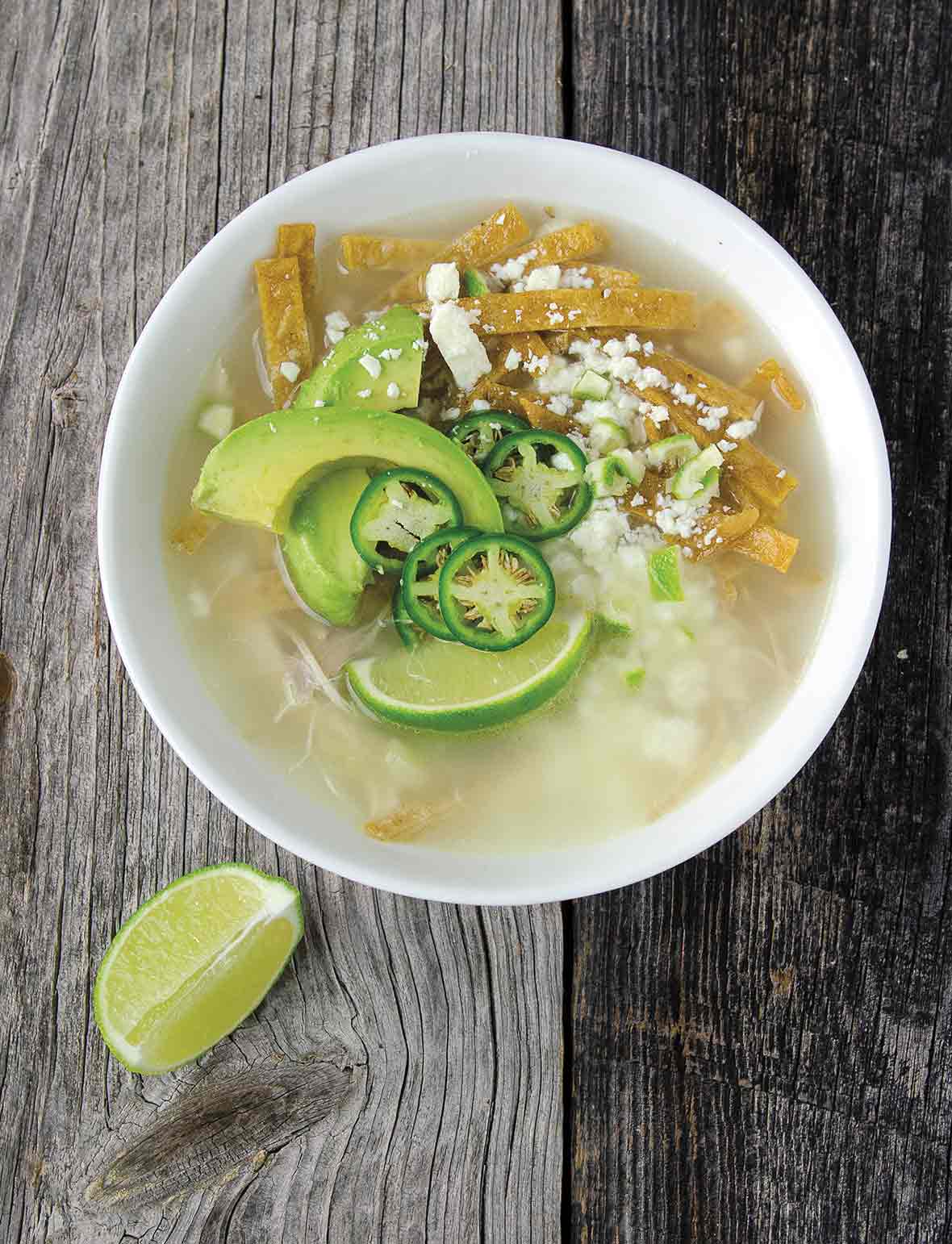 A white bowl filled with broth, avocado slices, lime wedges, cheese, and tortillas strips