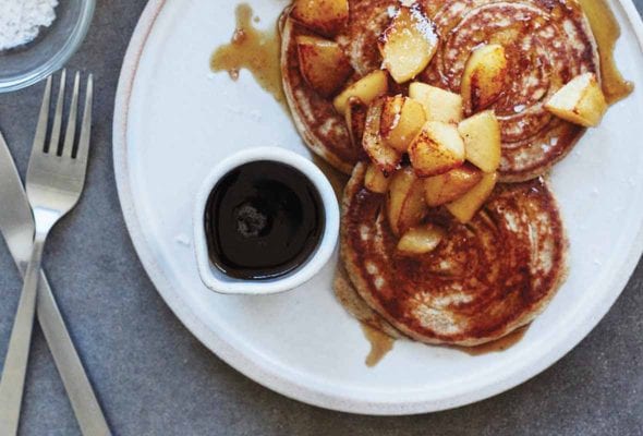 A plate with a small dish of maple syrup and 3 rye pancakes that are topped with maple syrup and pears.