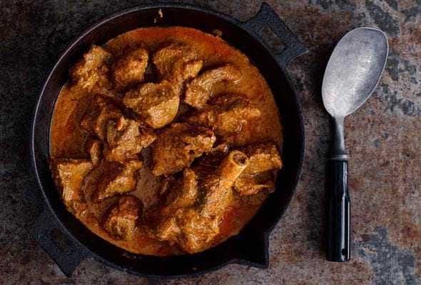 A bowl of lamb curry, known as laal maas with a silver spoon resting beside it.