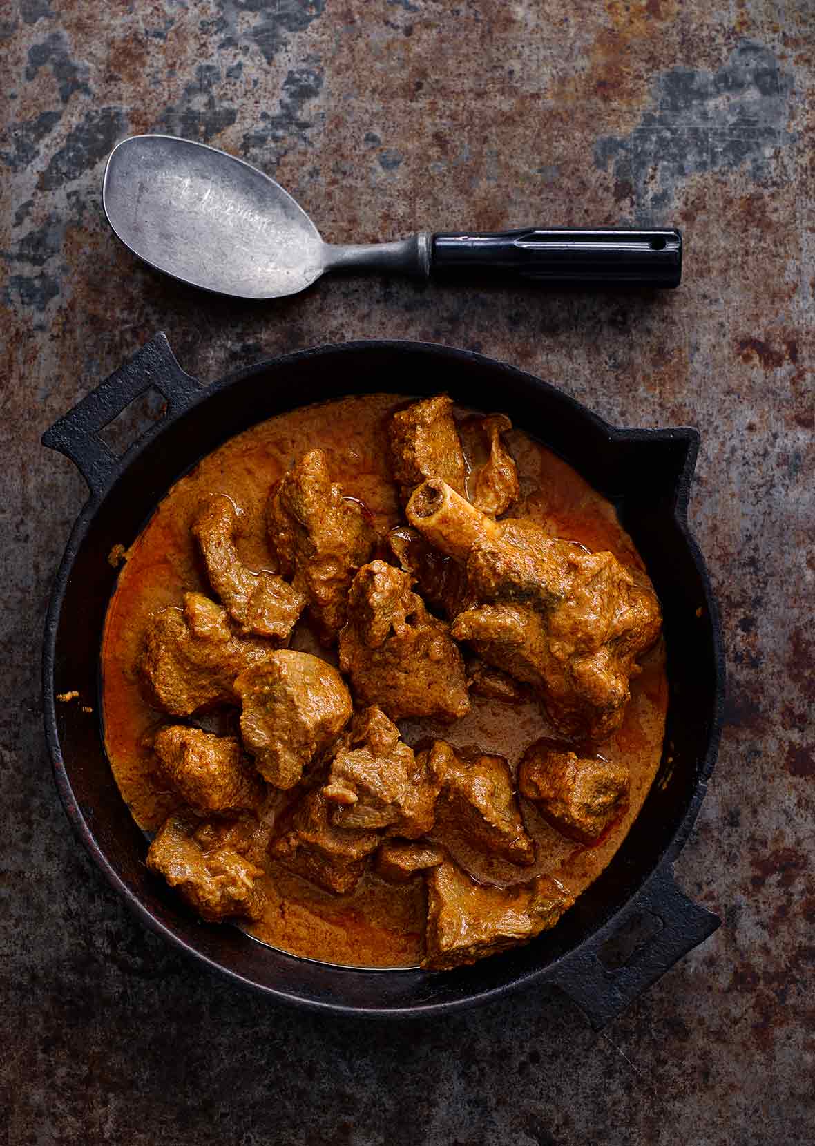 A bowl of lamb curry, known as laal maas with a silver spoon resting beside it.