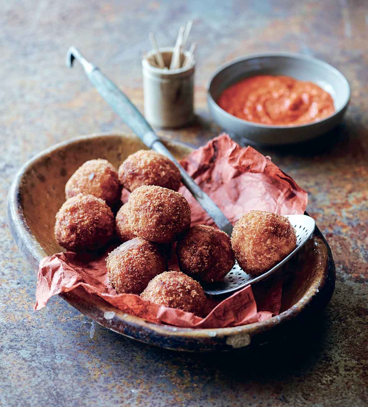 A large bowl filled with fried arancini balls and a small bowl of red pepper sauce on the side