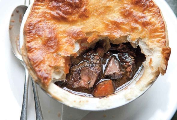 A venison pie with part of the crust missing on a white plate with a fork and spoon beside it.