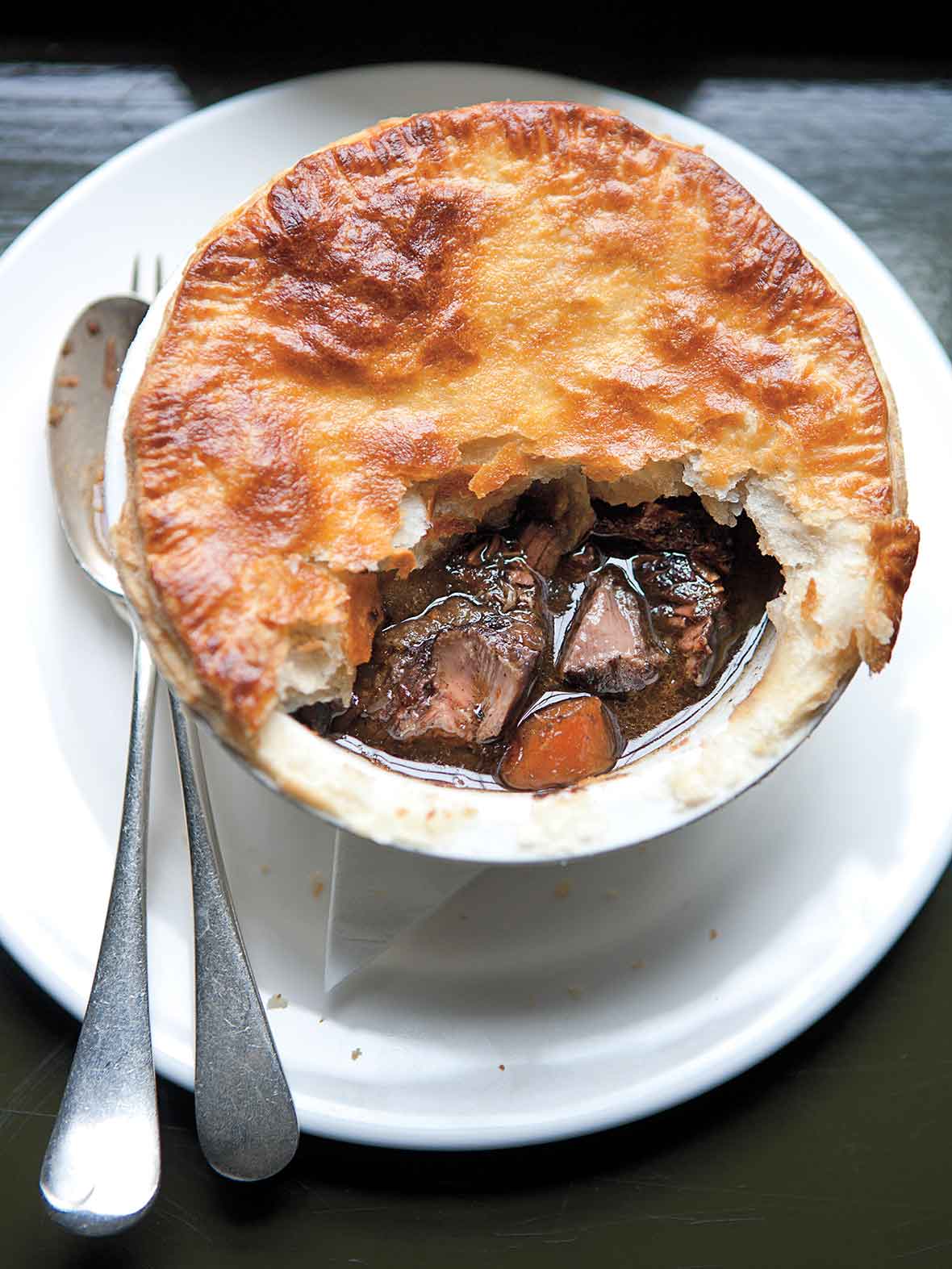 A venison pie with part of the crust missing on a white plate with a fork and spoon beside it.
