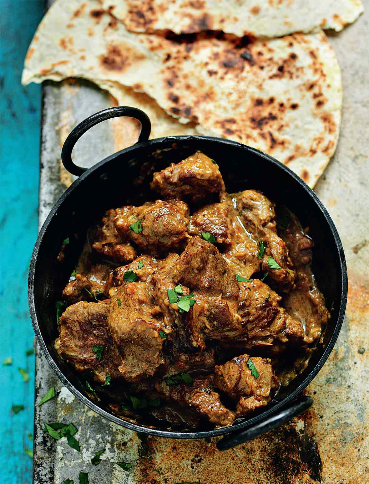 A black pot filled with chunks of beef Raj curry, topped with cilantro, nearby is roti Indian bread