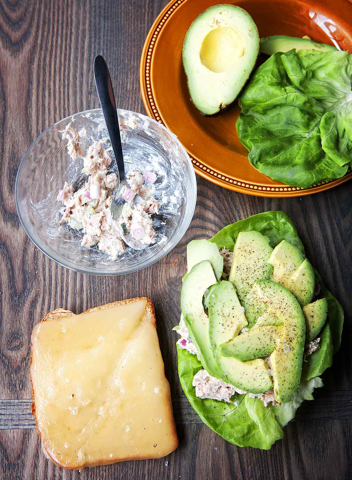 The makings of a Mediterranean tuna melt -- a slice of whole grain bread topped with gruyere cheese, a bowl with tuna salad, and a butter lettuce leaf topped with tuna salad, avocado, and pepper