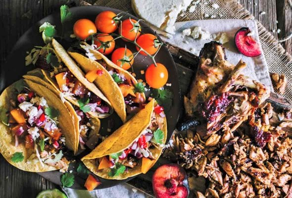 A black platter with several grilled chicken tacos with fruit salsa, a few cherry tomatoes, and a wooden board with cut chicken, halved plums, and queso fresco.
