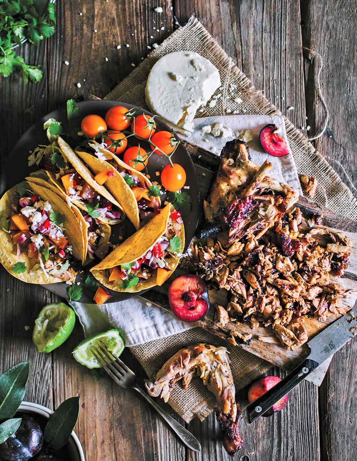 A black platter with several grilled chicken tacos with fruit salsa, a few cherry tomatoes, and a wooden board with cut chicken, halved plums, and queso fresco.