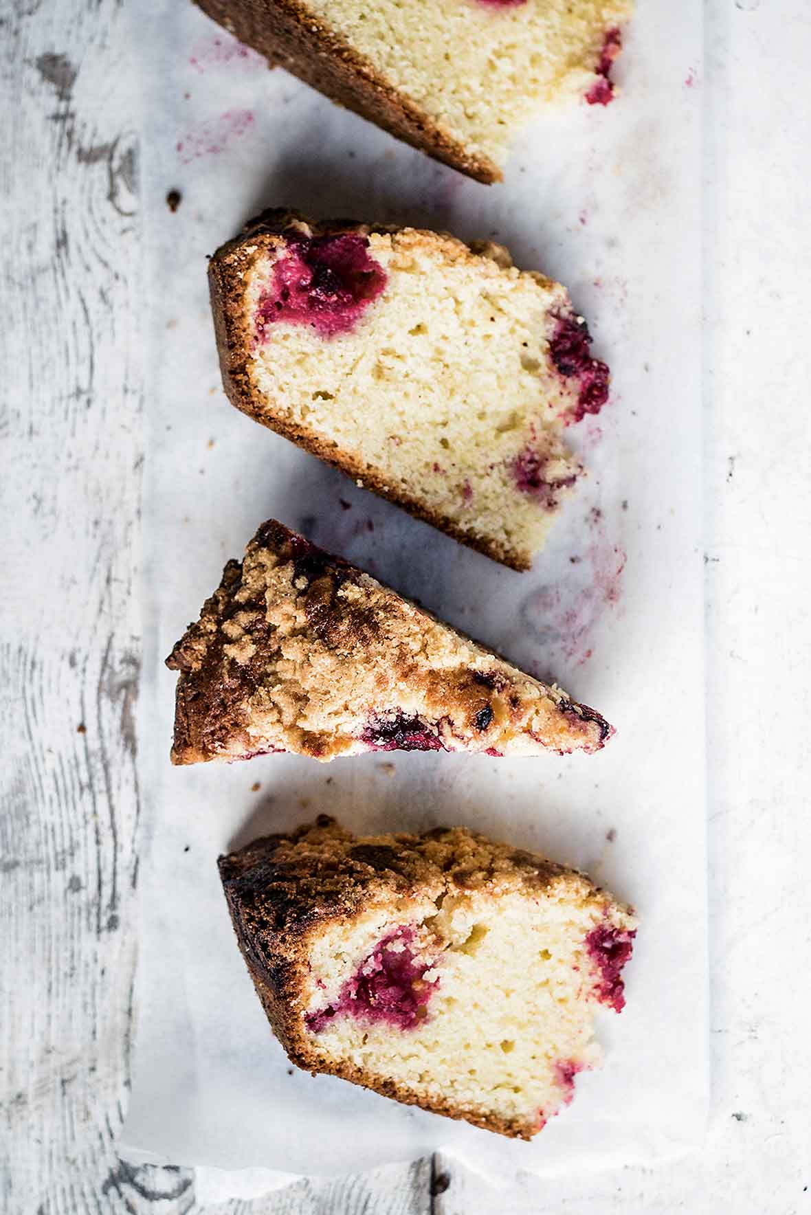 Four wedges of raspberry streusel coffee cake on a piece of parchment paper on a white wooden surface.
