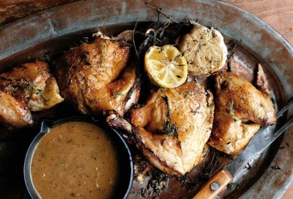 Pieces of roast chicken wth a small dish of pan gravy and a halved lemon and bulb of garlic on an oval metal tray.