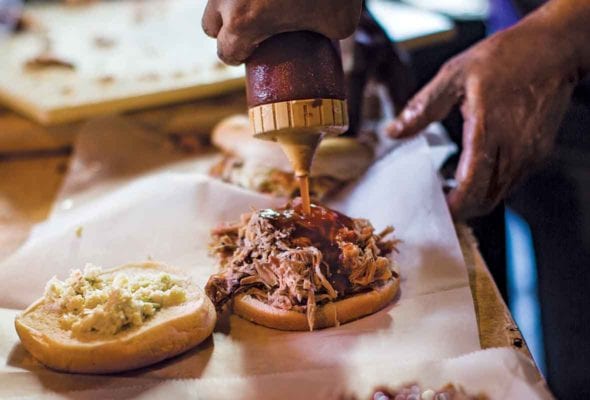 A person squeezing North Carolina barbecue sauce from a plastic bottle onto a sandwich.