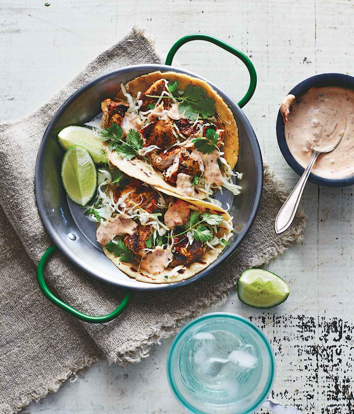 Two ancho fish tacos in a metal dish with lime wedges and chipotle mayo on the side.
