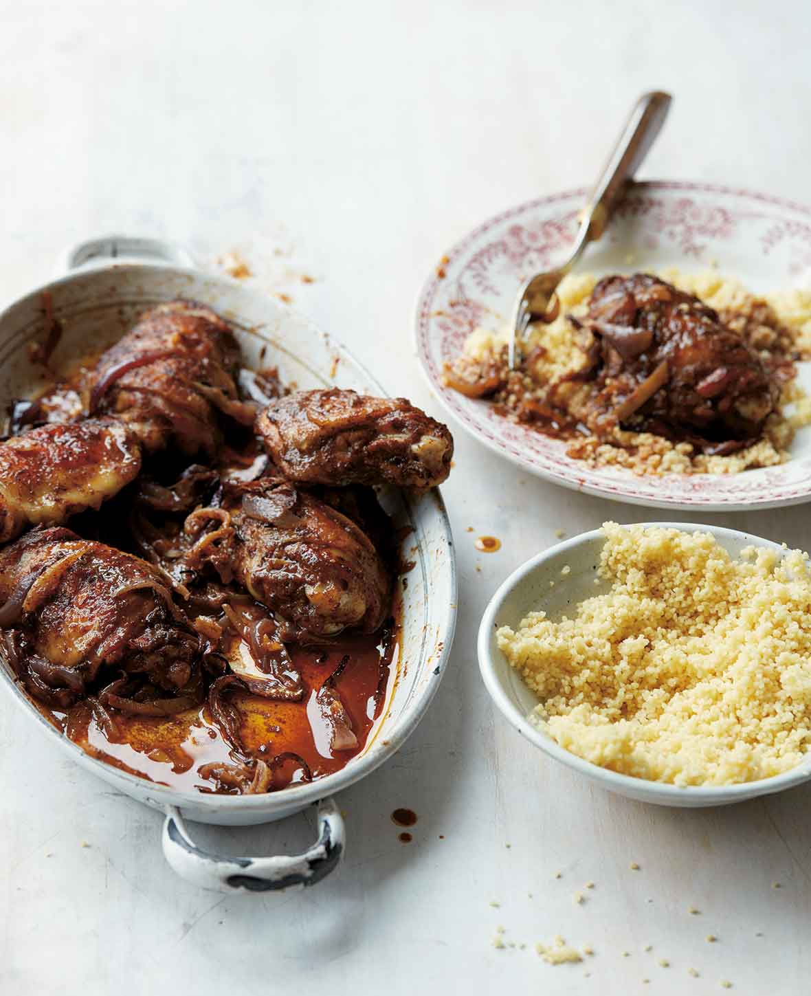 An oval dish filled with Baharat spiced chicken pieces and a bowl of couscous and plate of couscous and chicken on the side.