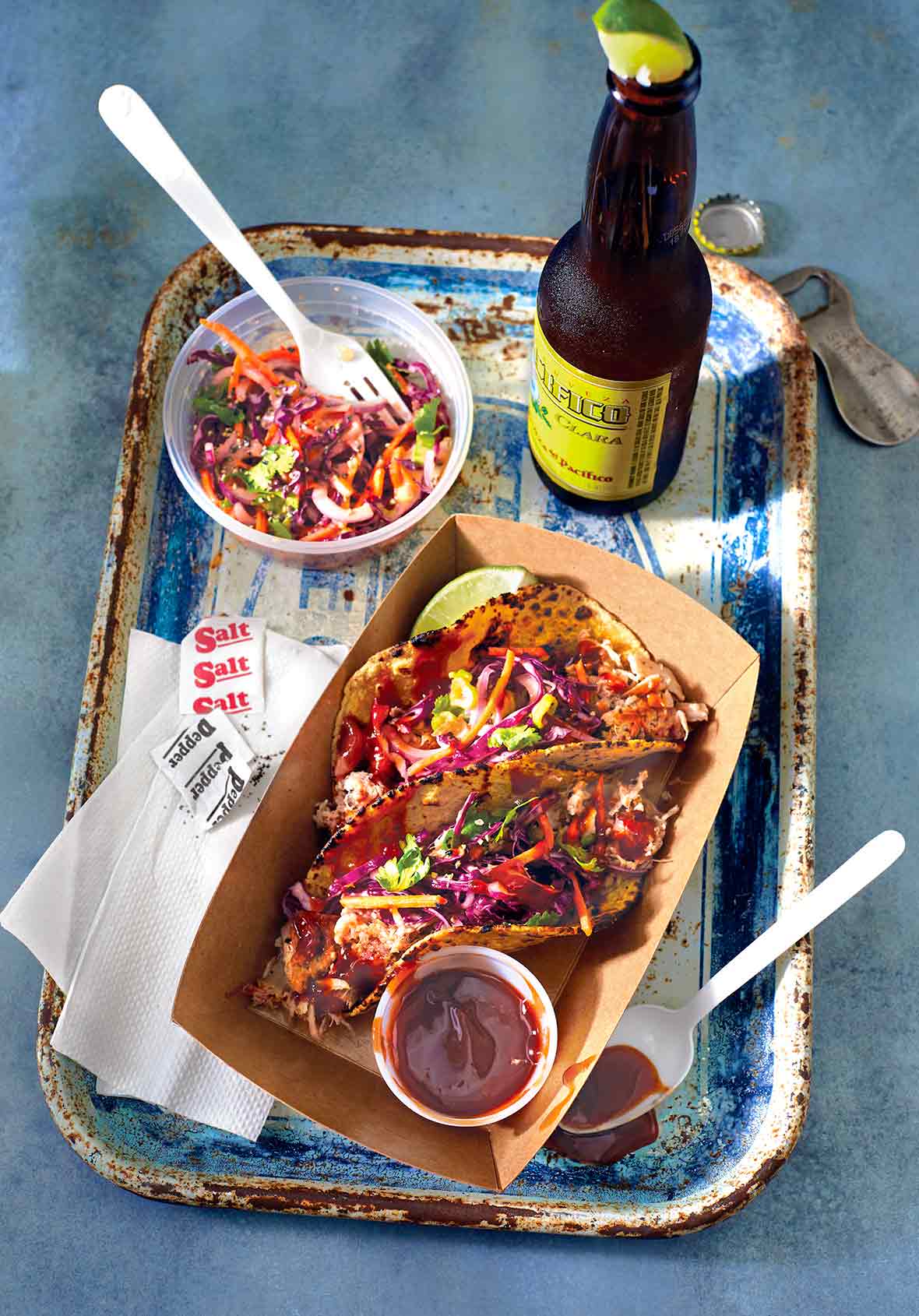 A cardboard tray with two pulled pork tacos topped with chipotle slaw and a bowl of slaw and bottle of beer on the side.